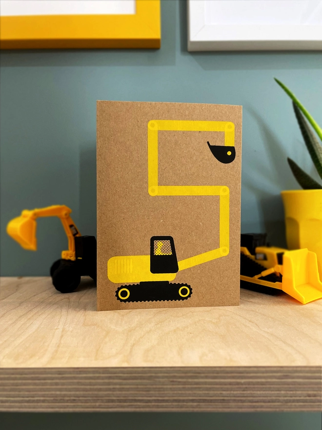 A yellow and black digger makes a number 5 shape, screenprinted on a Kraft brown card. Stood on a plywood shelf with 