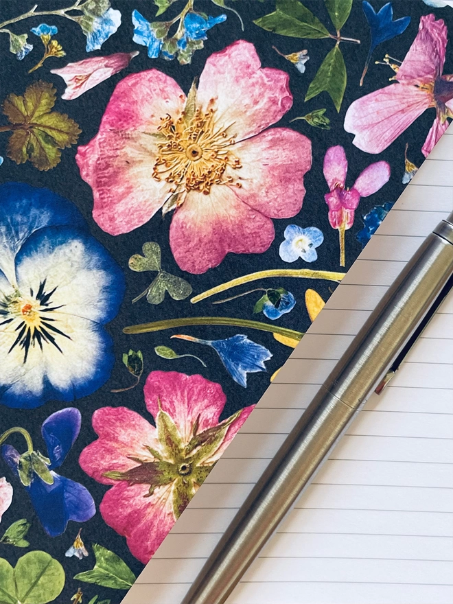 Close-Up of Beautiful Notebook with Decorative Pressed Flower Cover, Sitting on Open Notebook with Lined Pages, Silver Pen on Pages
