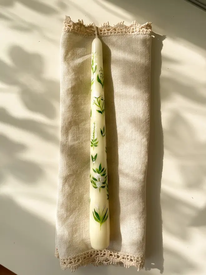 White Florals Dinner Candle, a white candle on a napkin on a plain surface. It's green floral design wraps around it. 