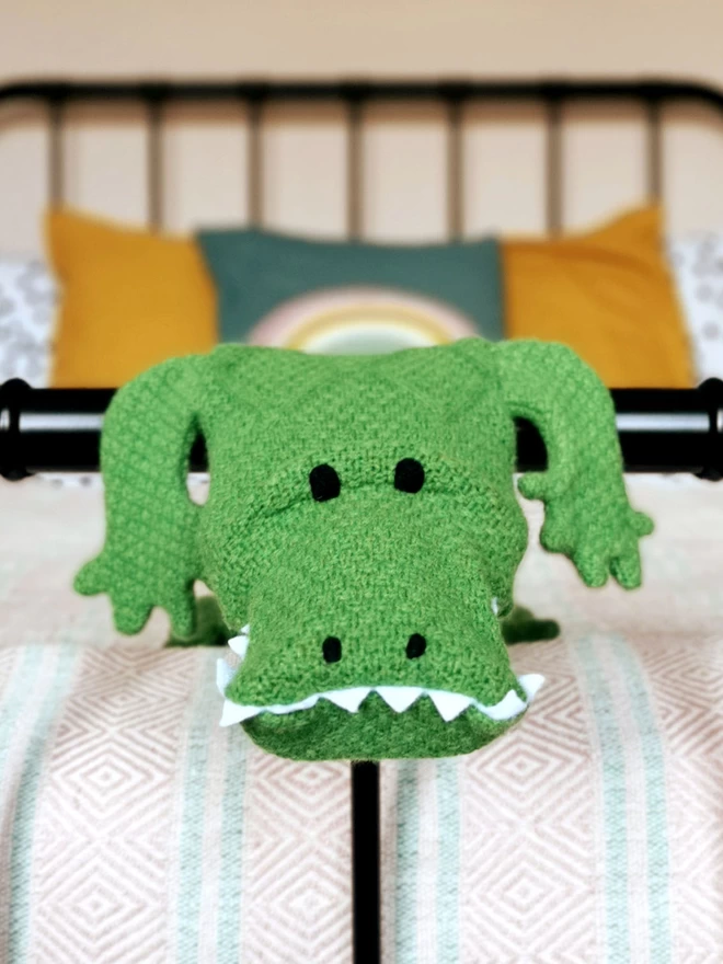 A close up of Claude the crocodile long hot water bottle face peeking over the end rail of a black metal framed double bed. He is looking cheekily straight into the camera.