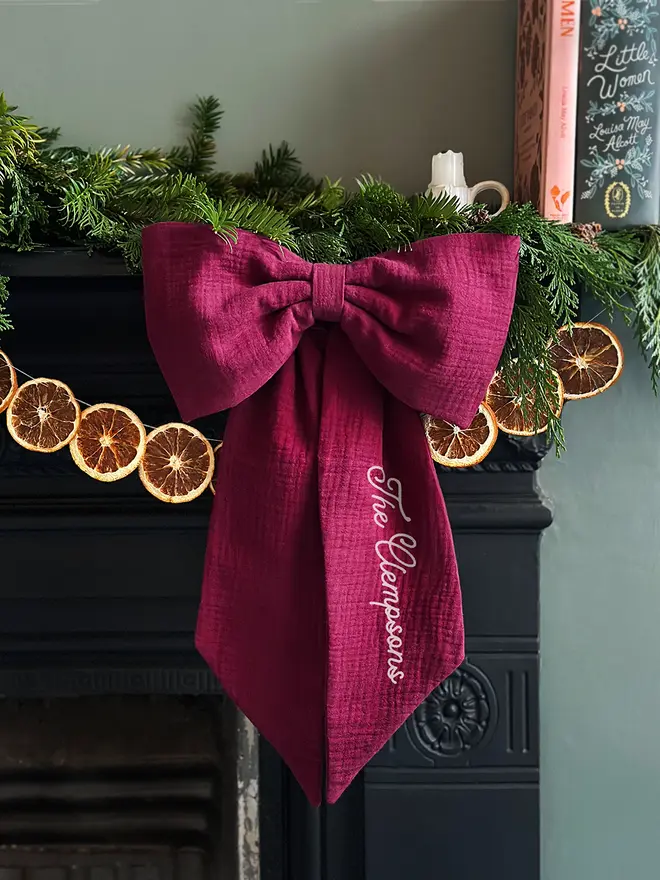 A handmade Christmas bow made from deep burgundy cotton with the words "The Clempsons" on one tail is hung from a black mantlepiece that is decorated for Christmas with greenery, orange slice garland, and candles.