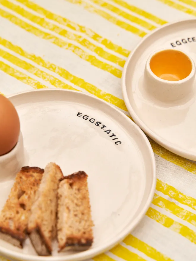 Egg And Soldier Plates, a plate with an integrated egg cup placed on a yellow striped surface with toast soldiers. The wording reads 'eggtastic' 