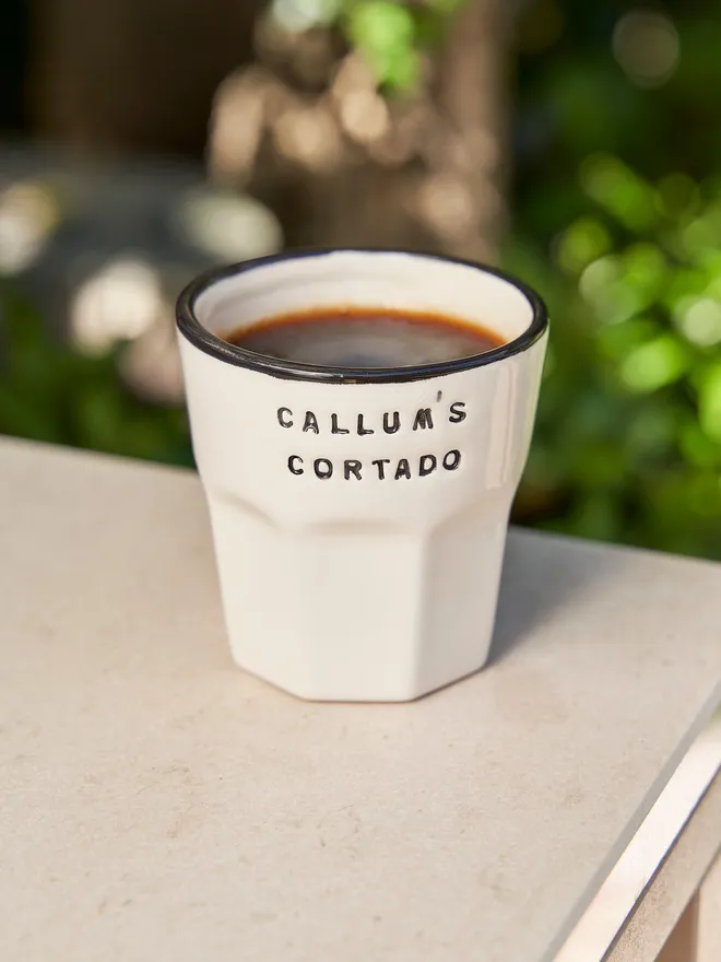 'Callum's Cortado' in a navy font stamped on a ceramic cup with a navy rim, on a table outside