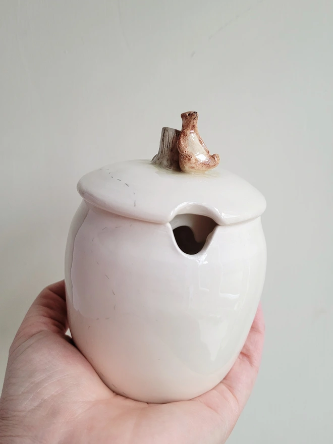 white pottery honey jar with a tiny brown bear siting on the lid being held in a hand