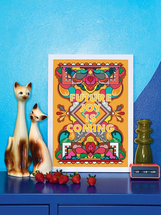 A vibrant portrait print on an orange background features Future Joy is Coming at the centre, surrounded by a rainbow coloured abstract design. The picture is in a white frame, against a turquoise and blue wall resting on a blue cabinet. Next to the picture are two cat ornaments, some ripe strawberries, a yellow glass vase and an orange Italian plastic calendar showing the date as ‘LU 10 LUG’.