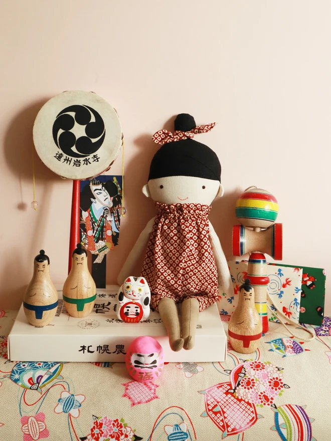 A handmade cloth doll with a light skin face, simple embroidered facial features, and black fabric hair styled with a matching patterned bow. The doll wears a red romper with a Japanese red shibori dots pattern.