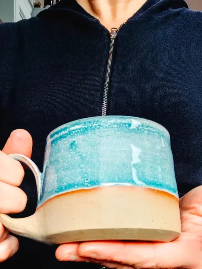 Light blue handmade ceramic mug held gently in two hands against a soft neutral background.