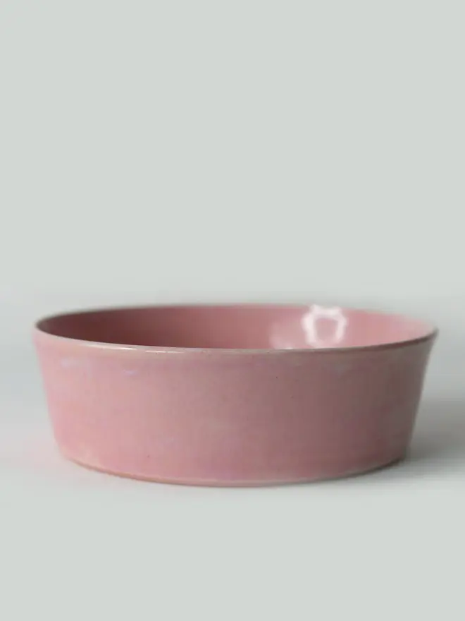 A single wide, shallow glazed ceramic pasta bowl in blush pink, photographed from a slight angle against a plain light grey background, showing the unglazed natural clay base.