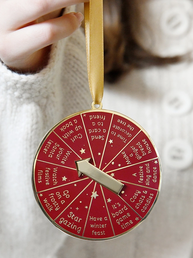 A deep red and gold Christmas decoration is being held by a gold ribbon. It has 12 segments, each one with a different Christmas activity idea, and a golden arrow in the centre.