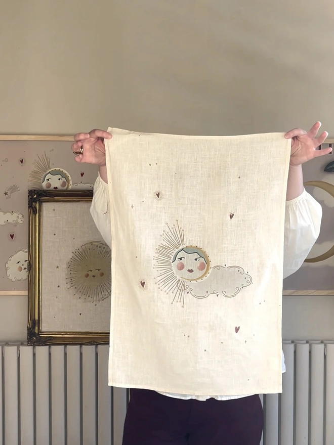 'Love Sun' Linen Tea Towel, a person holding up a linen tea towel with a sun motif on it. 