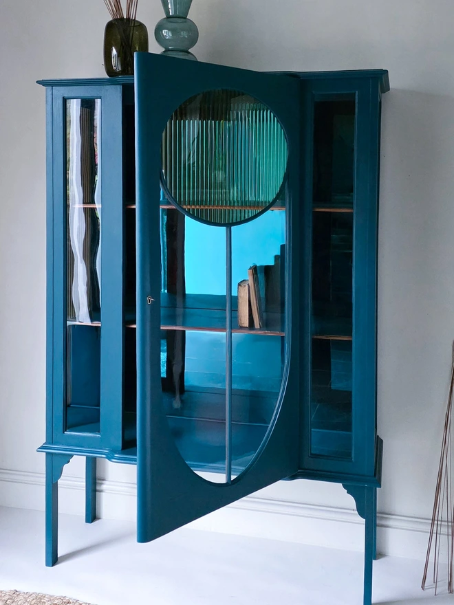Shows the door ajar of the teal and gold reeded glass vintage drinks cabinet, the interior has 2 wooden shelves in the matching colours each with the original wooden edging strip on show to add character. 