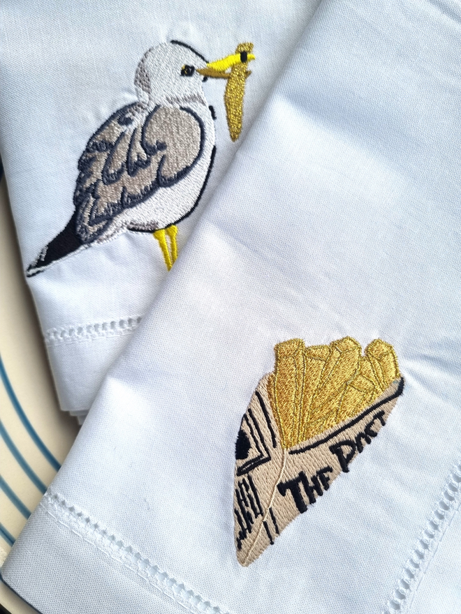 a set of two white cotton napkins with a seagull and chips embroidered 