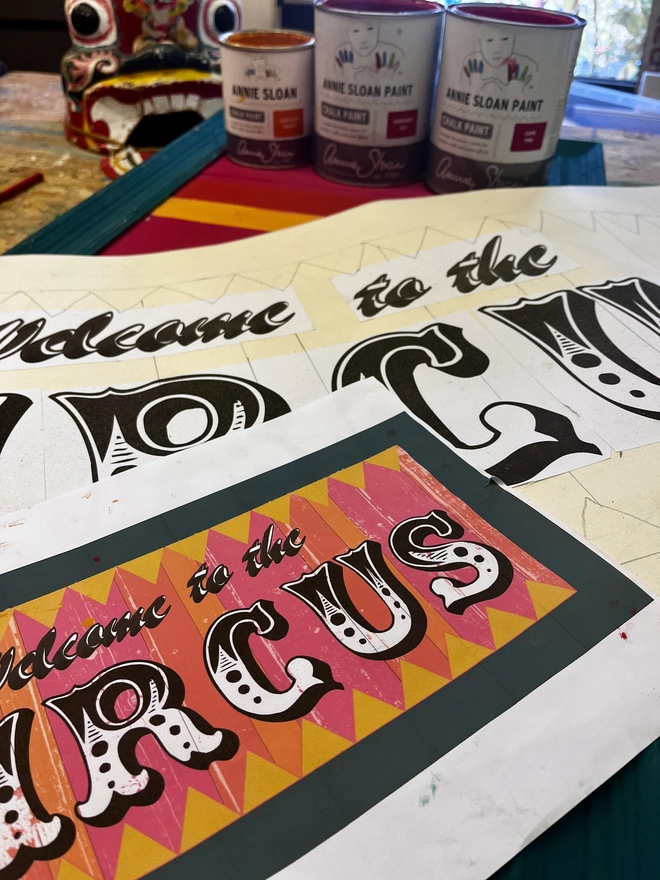 argent and sable workshop, showing design and painting process. welcome to the circus sign, with colourful stripes of pink, yellow and orange with a teal frame and zig zag pattern at the top and the bottom. surrounded by a teal frame
