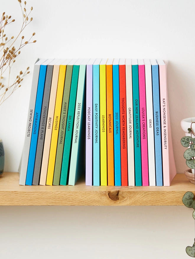 a rainbow of colourful notebooks stood up on a shelf with personalised text down the spine