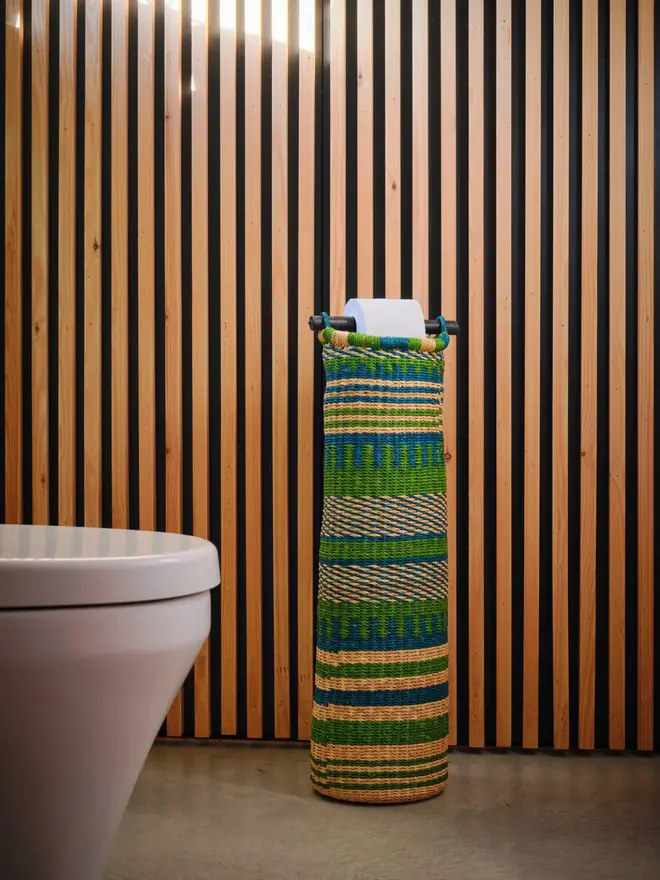 Ghanaian Hand Woven Tubular Toilet Roll Holder, a toilet roll holder sitting in a bathroom. It sits in front of a wooden backdrop. 
