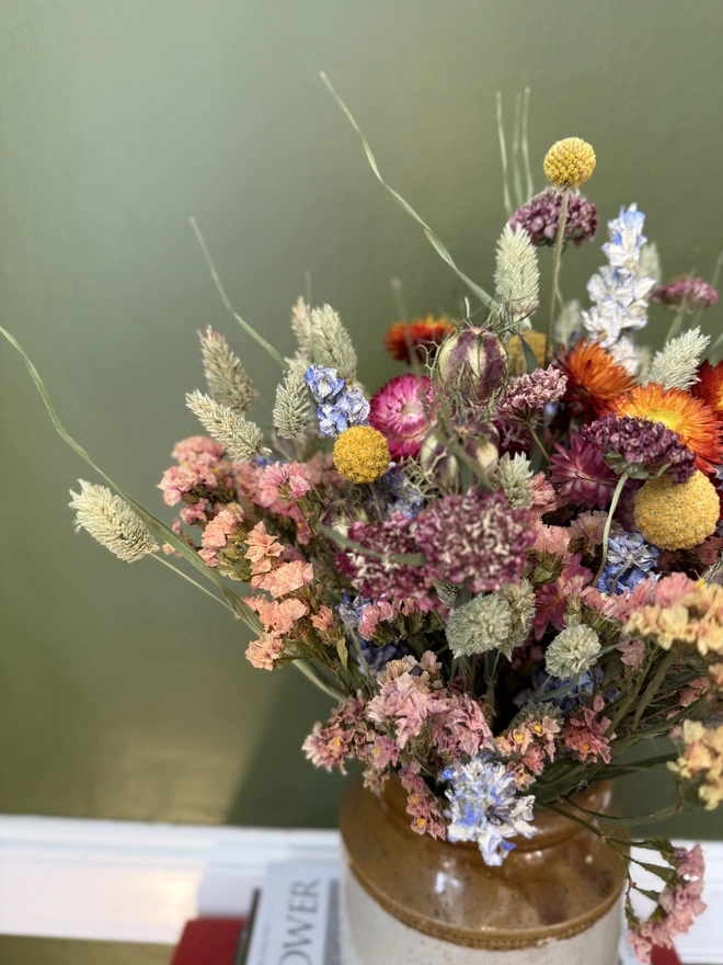 a dried flower bouquet of spring/summer colours in a victorian jam pot sitting on top of a stack of books and magazines against a green wall