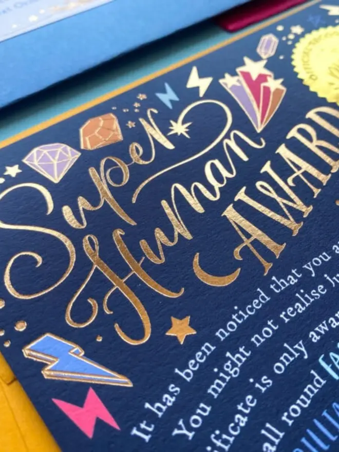 Close-up of the gold foil "Super Human Award" calligraphy on the navy certificate. Colourful gems, lightning bolts and shooting stars surround the lettering.