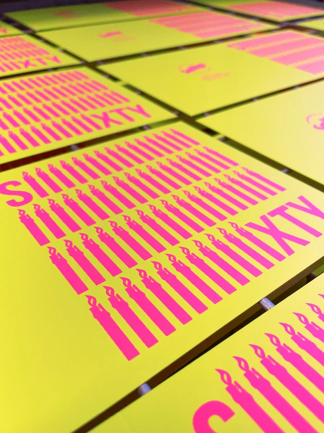 An angled shot of several unfolded screen printed Sixty birthday cards. On a drying rack. The word Sixty screenprinted in neon pink ink  across the bright yellow square cards. 