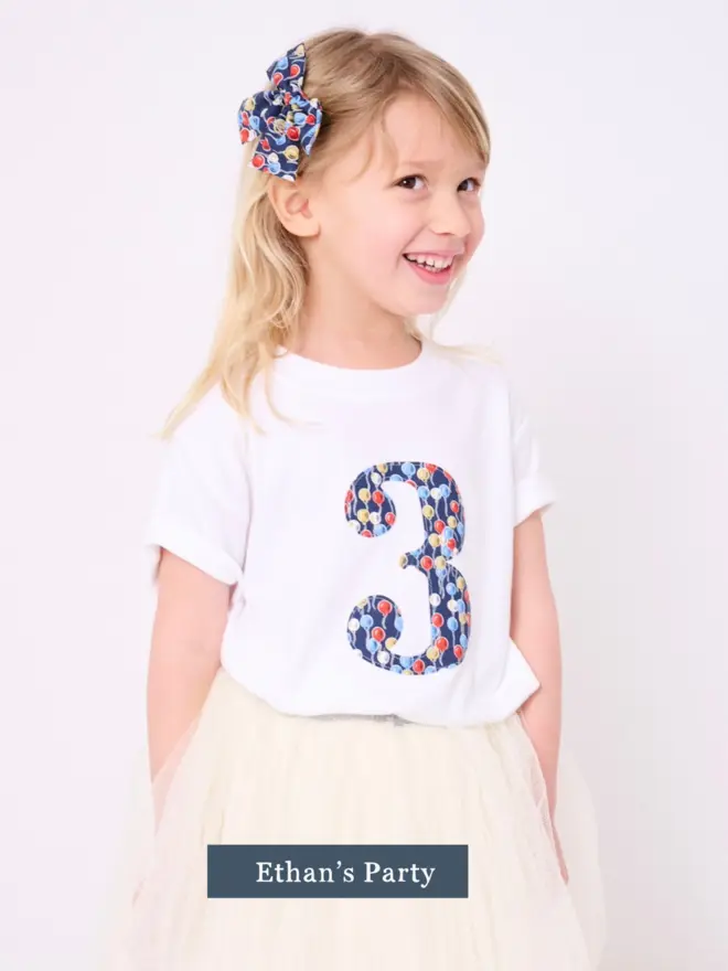 a 3 year old girl  wearing a white birthday t-shirt with a number 3 cut from liberty balloons print on the front