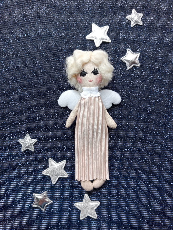 a tiny angel doll laying on a navy glittery backdrop with silver glittery stars placed around her. she is wearing a long pink gown. her hair is platinum white and softly curled