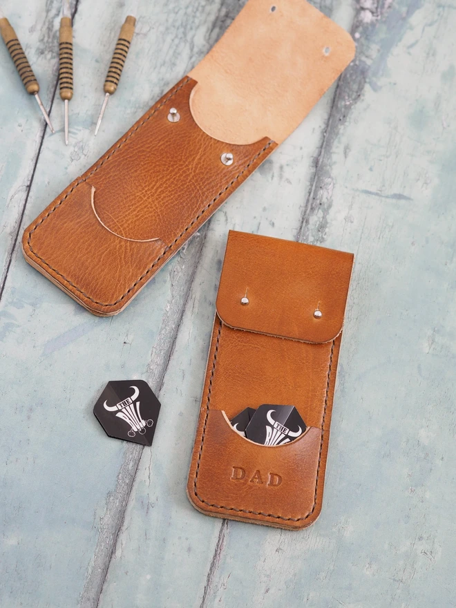 Personalised leather darts carry case. Two tan brown leather sleeves, one is personalised with DAD. Darts and flights sit around them.