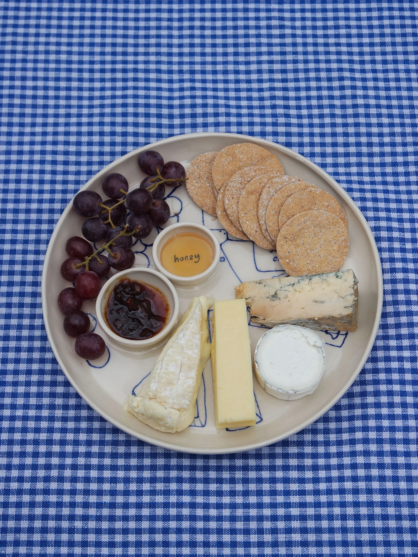 Dippy Cheeseboard Dinner Plate, a white dip plate sitting on a picnic blanket. It's covered in cheeses, crackers and grapes.