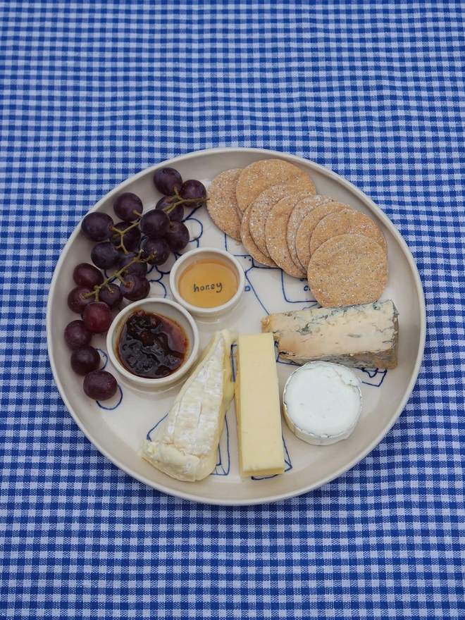 Dippy Cheeseboard Dinner Plate, a white dip plate sitting on a picnic blanket. It's covered in cheeses, crackers and grapes.