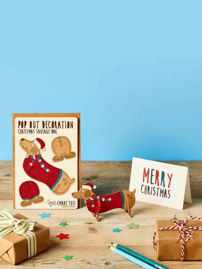 Pop out laser-cut Sausage Dog Christmas decoration and Merry Christmas card and brown kraft envelope on top of a wooden desk in front of a sky blue coloured background